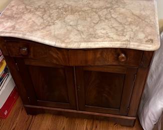 Marble-Top Vintage Wooden Cabinet — This vintage wooden cabinet features a distinctive beige marble top with natural veining and a matching marble backsplash. The cabinet is crafted from rich dark wood with a smooth finish, two paneled doors, and a single drawer with round wooden knobs. Measuring approximately 36 in height, 36 in width, and 18 in depth, this piece is perfect for adding classic charm to a dining room. The cabinet shows signs of wear consistent with age, including minor marble surface marks, but remains solid and functional. Its timeless design and durable materials make it a valuable addition to vintage furniture collections.