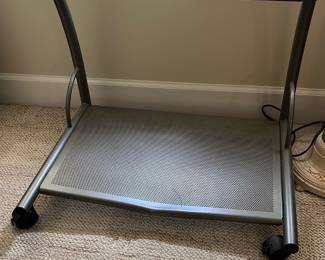 Metal Rolling Office Desk Cart — This metal rolling office desk cart provides practical storage and mobility for office equipment. Constructed from durable perforated metal with a silver-gray finish, it features two shelves supported by a sturdy tubular frame. Measuring approximately 24 inches high, 28 inches wide, and 18 inches deep, it is compact yet functional for small office spaces. The cart is in good condition with minor surface wear consistent with typical office use. Ideal for organizing printers, peripherals, or office supplies, this cart offers convenience and versatility.
