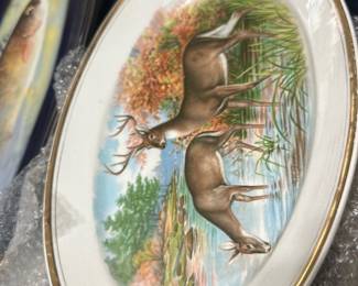 Decorative Deer Scenic Plate — Decorative porcelain plate featuring a painted deer scene with a gold rim and autumnal landscape. Plate shows detailed hand-painted style imagery of a buck and doe at water's edge with grasses and trees; gold trim around the edge. Measures approximately 11.5 inches diameter and is a lightweight decorative charger suitable for wall display or cabinet. Condition fair — crazing visible on the glaze and some surface wear to the backstamp but no visible cracks or large chips.