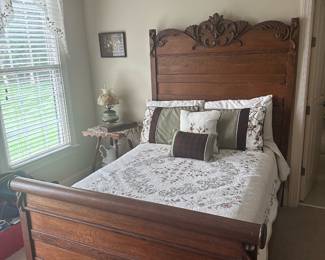 Carved Oak Full Bed Frame — Stately carved oak full-size bed frame with decorative crest and paneled footboard, photographed in the living room. Solid oak construction with a high headboard featuring floral scroll carving and a matching paneled footboard; warm medium-brown finish complements traditional decor. Overall footprint fits a full mattress; sturdy platform style suitable for standard box spring or slatted support. Good estate-condition with light surface wear and minor finish scuffs consistent with normal use; structurally sound and ready for re-use or refinishing.
