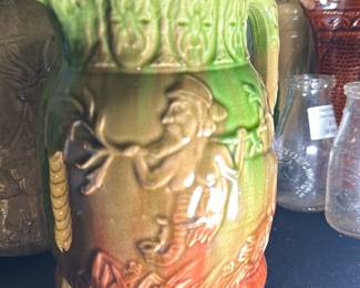 Green and Orange Ceramic Pitcher — This charming ceramic pitcher features a detailed relief design depicting a man playing a horn. The pitcher is glazed in vibrant green at the top, blending into warm orange at the base, with an intricate floral pattern adorning the rim. It measures approximately 8 inches in height and 6 inches in width, ideal for decorative display or functional use. The piece is in good condition with minor wear consistent with age, and no visible cracks or chips. Its unique design and warm colors make it an attractive addition to any vintage or rustic collection.
