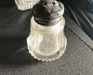 Vintage Glass Salt Shaker — This vintage glass salt shaker features a clear cut glass body with decorative ridges and a tarnished metal top with holes for dispensing. The compact shaker has a classic, timeless design suitable for any kitchen or dining table display. Measuring approximately 2.5 inches tall and 1.75 inches in diameter, it’s a collectible piece with visible signs of wear on the metal lid consistent with its age. The piece is in fair condition due to the tarnish but remains functional and an attractive addition to vintage kitchenware collections.