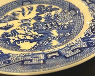 Semi China Blue Willow Decorative Plate — This Semi China decorative plate from England features the iconic Blue Willow pattern, showcasing a traditional Asian-inspired design with pagodas, trees, boats, and a bridge in vibrant blue on a cream background. The plate measures approximately 10.5 inches in diameter with a subtle raised rim and is crafted from semi-porcelain china. It exhibits light crazing and one visible chip on the rim, consistent with its age, adding to its vintage character. This collectible piece exemplifies mid-20th-century English ceramic artistry and makes a charming addition to any blue dishes collection or decorative display.