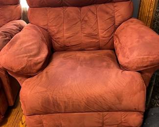 LaZBoy Recliner Chair — This rust red recliner chair offers plush comfort and a cozy seating experience for any living room. Upholstered in a soft, suede-like fabric, it features thick padded arms, a high cushioned back, and a matching pillow for added support. The chair measures approximately 42 inches in height, 38 inches in width, and 36 inches in depth, providing ample space for relaxation. It is in good condition with normal signs of wear on the seating area but remains fully functional. Perfect for lounging and adding a warm color accent to your living space.