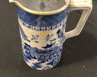 T. G. M. Co. Blue Willow Pitcher — This vintage T. G. M. Co. porcelain pitcher features the classic Blue Willow pattern in blue and white with traditional Asian-inspired scenic designs. It includes a metal hinged lid with a finial and a sturdy handle, perfect for serving beverages. The pitcher measures approximately 7.5 inches in height and 4.5 inches in width, making it a practical and decorative addition to any kitchen or dining collection. The piece is in good condition, displaying minor signs of wear consistent with age, and the metal lid shows some tarnishing. Marked on the base as "T.G.M.Co. Blue Willow," this collectible pitcher embodies the charm of English transferware.
