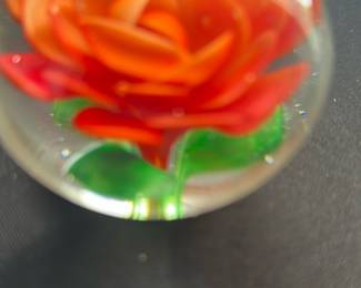 Red Rose Glass Paperweight — This charming glass paperweight features a vibrant red rose encapsulated within a clear spherical form. The intricate petals and green stem are skillfully crafted, showcasing a delicate and colorful floral design. Measuring approximately 3 inches in diameter, it is a lovely decorative accent suitable for any desk or display shelf. The piece is in good condition with no visible chips or cracks. Perfect for collectors of glass art or as a thoughtful gift item.
