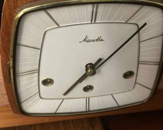 Mauthe Mid-Century Mantel Clock — This classic Mauthe mantel clock features a distinctive mid-century modern design with a wood case and brass accents. The clock face is white with an elegant geometric pattern and three winding holes, showcasing the brand's vintage German craftsmanship. Measuring approximately 6 in height, 10 in width, and 3 in length, it is a compact piece suitable for bedroom or living room decor. The clock is in good condition with some minor wear consistent with age and retains its original finish. A timeless collectible from the renowned Mauthe clockmakers, perfect for enthusiasts of vintage timepieces.