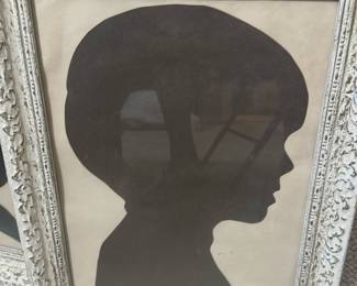 Framed Child Silhouette Portrait — Charming framed silhouette portrait of a child's profile in dark brown on a cream background, presented in an ornate distressed white frame. The piece appears to be paper cut or printed and is mounted behind glass with decorative molded frame detailing. Good size for wall or tabletop display in a nursery, hallway, or gallery wall; frame shows light surface wear consistent with age. Overall condition is good with minor scuffs to frame and slight toning to the paper; ready to hang or display.