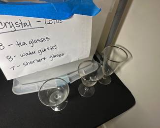 Crystal Lotus Glassware Set — This crystal Lotus glassware set includes tea glasses, water glasses, and sherbet glasses as indicated by the seller's notes. The collection features delicate stems and clear bowl shapes suitable for various drinks, making it an elegant addition to any table setting. There are 8 tea glasses, 8 water glasses, and 7 sherbet glasses stored in a labeled plastic container in the bonus up room. The glasses appear to be in good condition with no visible chips or cracks. Perfect for formal dining or special occasions.