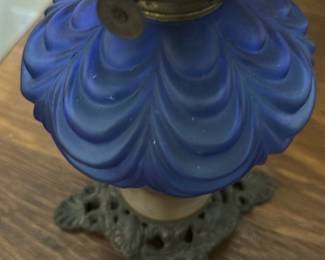 Vintage Blue Glass Oil Lamp — This vintage oil lamp features a stunning cobalt blue glass chimney and reservoir with a decorative swirl pattern. The lamp includes an ornate metal base with intricate detailing, adding to its antique charm. Standing approximately 15 inches tall and 6 inches wide, it makes a beautiful accent piece for any bedroom or living space. The lamp is in good condition with some minor wear consistent with age, but the glass and metal components are intact. Its classic design evokes a nostalgic ambiance ideal for collectors or vintage decor enthusiasts.