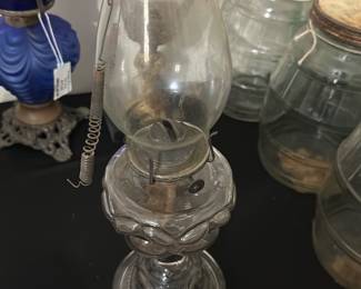 Vintage Glass Oil Lamp — This vintage glass oil lamp features a classic design with a clear glass chimney and an ornate glass base. The lamp includes the original wick holder and a metal spring mechanism for adjusting the wick. Measuring approximately 12 inches tall and 5 inches wide at its base, it offers both decorative appeal and functional use. The lamp is in good condition with some signs of age, including minor cloudiness on the glass and surface wear consistent with vintage oil lamps. Ideal for collectors or as a unique home decor accent.