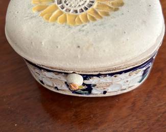 Ceramic Trinket Box — This charming Hughes Treasures ceramic trinket box features a sunflower design on the lid and a decorative floral band around its sides. The box has an oval shape and a hinged lid connected by leather cords. Measuring approximately 2.5 inches tall, 5 inches wide, and 4 inches deep, it provides a lovely small storage solution. The item is in good vintage condition with minor surface wear indicative of age. This collectible ceramic piece is perfect for storing jewelry or keepsakes, adding a rustic decorative touch to any room.