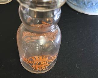 Ezell Purity Dairies Mackie Milk Bottle — This vintage Ezell Purity Dairies Mackie glass milk bottle is a charming collectible from mid-20th century dairy delivery. The clear glass bottle features bright orange vintage branding with the company name and contact dial number. Measuring approximately 6 inches in height and 3.25 inches in width, this sturdy bottle is a great display piece or addition to a dairy memorabilia collection. It shows signs of use consistent with age but remains intact without chips or cracks. A nostalgic representation of classic milk delivery, this bottle brings a touch of Americana to any collection.