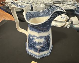 Adams & Co. Blue Transferware Pitcher — This elegant Adams & Co. blue transferware pitcher features a picturesque landscape scene framed by intricate border patterns. Crafted from fine ceramic with a distinctive octagonal shape, this pitcher showcases classic 19th-century English ceramic artistry. Standing approximately 8 inches tall and 6 inches wide at its base, it is a perfect decorative or functional piece. The pitcher is in good vintage condition with typical crazing and minor wear consistent with its age. Marked with the Adams & Co. Athens pattern and model number 6978, it represents collectible English transferware from a respected maker.