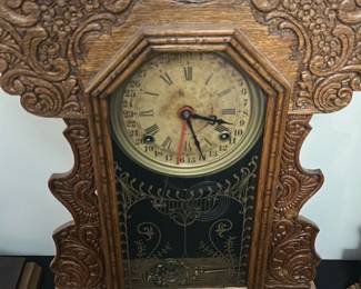 Antique Carved Oak Mantel Clock — This antique carved oak mantel clock features intricate woodwork with ornate floral and geometric patterns surrounding a glass face. The dial displays Roman numerals alongside a 31-day calendar, adding both stylistic charm and functionality. Measuring approximately 14 inches tall, 10 inches wide, and 4 inches deep, it is a decorative timepiece ideal for traditional home decor. The clock shows signs of age including patina and slight discoloration on the dial but remains an attractive vintage collectible. Perfect for collectors of antique clocks or those looking to enhance a vintage-styled bedroom space.