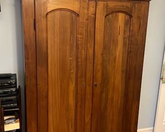 Walnut Armoire with Carved Crest — This substantial walnut armoire features a decorative carved crest and paneled double doors, offering generous storage with shelves and a hanging section. Constructed from solid walnut (visible grain and age patina) with removable interior shelves on the right and a hanging rod on the left, it has a classic late-19th/early-20th-century wardrobe look. Overall dimensions approximate a tall bedroom wardrobe suitable for master bedroom placement (measurements estimated below); heavy solid-wood construction will require two people to move. Good vintage condition with normal surface wear, small scuffs to shelf edges, and intact structural joinery; hardware and finish show age-appropriate patina.