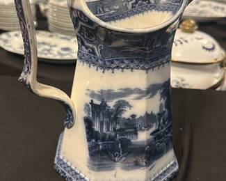 Adams & Co. Blue Transferware Pitcher — This elegant Adams & Co. blue transferware pitcher features a picturesque landscape scene framed by intricate border patterns. Crafted from fine ceramic with a distinctive octagonal shape, this pitcher showcases classic 19th-century English ceramic artistry. Standing approximately 8 inches tall and 6 inches wide at its base, it is a perfect decorative or functional piece. The pitcher is in good vintage condition with typical crazing and minor wear consistent with its age. Marked with the Adams & Co. Athens pattern and model number 6978, it represents collectible English transferware from a respected maker.