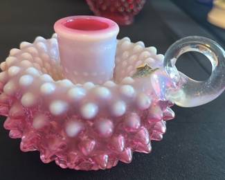 Fenton Cranberry Opalescent Candleholder — This lovely Fenton cranberry opalescent hobnail candleholder features a distinctive clear glass loop handle and a textured hobnail pattern for a vintage charm. Crafted from hand-blown glass with a white opalescent finish fading into cranberry, it serves as an elegant accent for any room. Measuring approximately 3 inches in height, 5 inches in width, and 4 inches in length, it is a perfect size for decorative use or collecting. The piece is in excellent condition showing no chips or cracks, retaining the original Fenton label that attests to its authenticity. This candleholder is a classic example of mid-20th century American art glass from the renowned Fenton Art Glass Company.