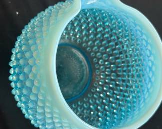 Blue Hobnail Milk Glass Pitcher — This charming blue hobnail milk glass pitcher features a textured bubble design with a clear twisted handle for easy pouring. The pitcher exhibits a beautiful opaline blue color with classic vintage appeal. Measuring approximately 5.5 inches tall and 6 inches wide including the handle, it makes an elegant accent for any tabletop or display. The piece is in good condition with no visible chips or cracks, showing minor wear consistent with vintage glassware. Its distinctive hobnail pattern and opaline glass make it a collectible item for vintage glass enthusiasts.
