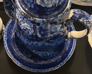 Blue Transferware Teapot and Saucer — This charming blue transferware teapot and matching saucer feature intricate floral and scenic designs characteristic of classic blue and white pottery. The teapot has a detailed curved handle, spout, and a domed lid with a finial, paired with a saucer showcasing a scenic rural estate design. Measuring approximately 6.25 inches in height and 9 inches in width including the handle and spout, this set is suitable for display or serving. The item is in good condition with some typical wear to the glaze that adds to its vintage appeal. This set represents timeless traditional ceramic craftsmanship, often found in collectible blue dishes room collections.