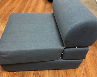 Blue Upholstered fold out  Chair — This versatile blue upholstered chair is designed for comfortable seating, ideal for basement or casual living areas. Featuring a simple block form with a rounded backrest, the chair is covered in a durable blue fabric. Its compact size makes it suitable for smaller spaces or as additional seating. The chair appears to be in good condition with minor wear from normal use. Perfect for someone seeking functional and cozy furniture for informal settings.