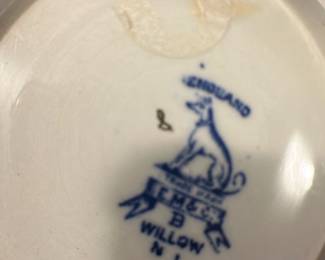 T. G. M. Co. Blue Willow Pitcher — This vintage T. G. M. Co. porcelain pitcher features the classic Blue Willow pattern in blue and white with traditional Asian-inspired scenic designs. It includes a metal hinged lid with a finial and a sturdy handle, perfect for serving beverages. The pitcher measures approximately 7.5 inches in height and 4.5 inches in width, making it a practical and decorative addition to any kitchen or dining collection. The piece is in good condition, displaying minor signs of wear consistent with age, and the metal lid shows some tarnishing. Marked on the base as "T.G.M.Co. Blue Willow," this collectible pitcher embodies the charm of English transferware.