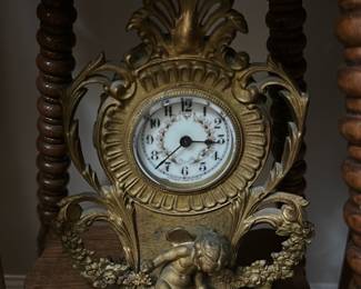 Antique Gold Cherub Mantel Clock — This charming antique mantel clock features an ornate gold-tone design with intricately detailed cherub figure and floral garland accents. The clock face has vintage black numerals and elegant minute markings set on a white background with a decorative laurel wreath. Measuring approximately 9 inches in height, 7 inches in width, and 3 inches in depth, it is perfect for display on a mantel or shelf. The clock is in fair condition with some tarnishing and wear consistent with its age, lending it added character. A beautiful decorative piece representing classic Victorian style and craftsmanship.