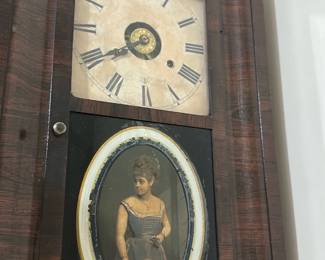 Victorian Walnut Wall Clock — Victorian-era walnut wall clock with painted dial and reverse-painted lower tablet depicting a woman in period dress. The piece features a rectangular rosewood-grain case, Roman numeral face, and original metal hands; sold from the bedroom up room. Measures approximately 28 inches tall by 16 inches wide and 4 inches deep, making it a compact decorative wall clock for a hall or parlor. Clock shows age-appropriate wear: crazing and staining on the dial, minor chips and surface wear to the painted lower glass, and light scuffs to the veneer. Mechanism condition unknown — likely needs servicing for reliable timekeeping but displays beautifully as period wall decor.