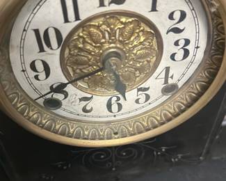 Antique Black Resin Mantel Clock — This antique black mantel clock features elegant brass detailing and ornate columns with a classic design. The clock face displays bold black numerals around a gold embossed center, enclosed in a brass bezel. Measuring approximately 12 inches tall, 16 inches wide, and 6 inches deep, it is a substantial piece ideal for display on a mantel or shelf. The clock shows some wear consistent with age, including minor chipping and surface scratches, but remains a charming vintage collectible. Its Victorian-era styling and craftsmanship make it a distinctive addition for antique enthusiasts.
