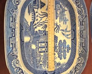 Blue Willow Platter — This vintage Semi China blue willow platter features the classic blue and white chinoiserie pattern with intricate pagodas, trees, and birds. Crafted from semi-porcelain china, this platter measures approximately 14 inches in length and exhibits charming age-related crazing and a minor chip near the edge. It is a perfect decorative or functional serving piece, embodying traditional English china artistry. The piece is in fair condition due to visible wear but retains its vintage character and collectible value.