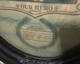 Our Hero Oval Memorial Print — Striking oval framed memorial print titled 'Our Hero' featuring a young serviceman portrait with patriotic motifs and laurel wreath. The piece has a carved black wooden oval frame (scuffs and nicks visible) with printed banners, flags and an eagle motif surrounding the central image. Measures approximately 20 in tall by 16 in wide and is a lightweight wall piece suitable for a study or gallery wall. Condition is fair with surface wear to frame, some glass glare and age-toning to the paper; a desirable patriotic memorial print for collectors of militaria and folk photography.