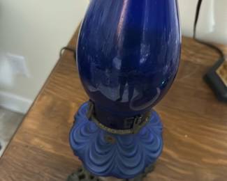 Vintage Blue Glass Oil Lamp — This vintage oil lamp features a stunning cobalt blue glass chimney and reservoir with a decorative swirl pattern. The lamp includes an ornate metal base with intricate detailing, adding to its antique charm. Standing approximately 15 inches tall and 6 inches wide, it makes a beautiful accent piece for any bedroom or living space. The lamp is in good condition with some minor wear consistent with age, but the glass and metal components are intact. Its classic design evokes a nostalgic ambiance ideal for collectors or vintage decor enthusiasts.