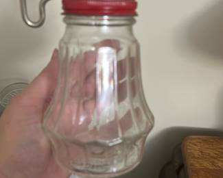 Vintage Glass Syrup Dispenser with Red L — This vintage glass syrup dispenser features a distinctive fluted design with a clear body and a bright red metal lid equipped with a unique metal handle lever. The glass construction suggests durability and easy cleaning, while the size makes it ideal for countertop use in a kitchen or dining area. The dispenser measures approximately 5.5 inches in height, 3.5 inches in width, and 3.5 inches in depth, making it a compact yet practical item. It is in good condition with minor wear consistent with age, and the lid mechanism functions properly. This classic kitchen accessory offers nostalgic charm and practical functionality for everyday use.