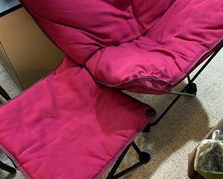 Red Folding Lounge Chair w/ Ottoman — This red folding lounge chair features a padded cushion over a metal frame, providing comfortable seating ideal for indoor or outdoor use. The chair folds flat for easy storage and portability, making it great for casual seating or small spaces. It measures approximately 36 inches high, 24 inches wide, and 30 inches deep, fitting nicely in a bonus room or living area. The chair is in good condition with minor signs of wear on the fabric but remains fully functional. Lightweight and versatile, this chair is perfect for layering extra seating in any living space.