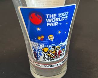 1982 World's Fair Souvenir Glass — This collectible glass features a colorful design commemorating the 1982 World's Fair held in Knoxville, Tennessee. The clear glass displays iconic fair imagery on a blue background with detailed characters and balloons. It measures approximately 4.5 inches tall and 3 inches in diameter at the top, making it a perfect vintage souvenir or display piece. The glass is in good condition with some minor age-related wear but no chips or cracks. A nostalgic artifact from an important cultural event, ideal for collectors of World's Fair memorabilia.