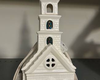 White Ceramic Church Lighted House — Charming white ceramic church-shaped light with molded brick and steeple details and faux stained-glass window inserts. The piece is a plug-in decorative house likely used as a holiday or mantel accent and features an electrical cord for internal illumination. Measures approximately 12 inches tall, 6 inches wide and 6 inches deep — a compact, easy-to-place decoration for shelves or tabletops. In good condition with no visible chips or cracks; cord appears intact though functionality was not tested.