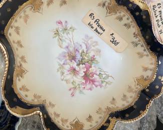 RS Prussia Blue Floral Bowl — This RS Prussia bowl features a classic blue border with intricate gold detailing and a central hand-painted floral motif in soft pinks and greens. The bowl showcases ornate scalloped edges with raised beadwork in gold, highlighting its fine craftsmanship. Measuring approximately 8 inches in diameter, this decorative porcelain piece is perfect as a display item or for elegant serving. It is in good condition with minor wear consistent with age and no visible chips or cracks. RS Prussia is renowned for quality German porcelain, making this bowl a valuable collectible from the early 20th century.
