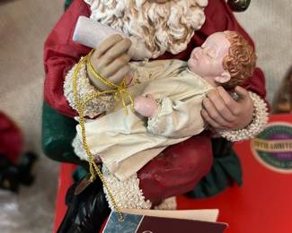 Clothtique Santa with Baby Figurine — Clothtique Santa holding and feeding a sleeping baby in a cream gown — a classic holiday collectible from the Clothtique Santas Possible Dreams collection. Santa wears a red suit with textured cream trim, painted resin face and hands, cloth clothing details, and a bubbled jingle bell on his hat; a tag reading "Clothtique Santas Possible Dreams" is attached. Measures approximately 10 inches tall and is a tabletop holiday accent perfect for mantel or shelf display. In good condition with light shelf wear visible on fabric trim and paint; appears complete with original tag and collectible box shown in photos.
