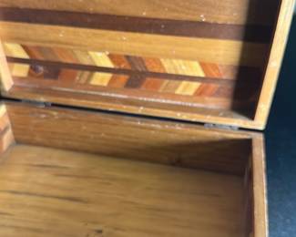 Wood Inlay Jewelry Box — This wood inlay jewelry box features a beautiful geometric pattern crafted from various wood tones, creating a distinctive decorative effect. The box is rectangular with a hinged lid and a small wooden latch for secure closure. Measuring approximately 3 inches tall, 8 inches wide, and 5 inches deep, it is a practical size for storing small collectibles or keepsakes. The box shows some surface wear and scratches consistent with age but remains sturdy and functional. Its handcrafted design and natural wood materials make it a charming vintage decorative piece suitable for a bonus up room or dresser.