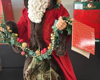 Red Hooded Santa Figurine — Festive red-hooded Santa figurine dressed in a red velvet-like cloak with faux-fur trim and holding a decorative berry and greenery garland. The figure features a detailed molded face and sculpted white beard with a textured gold and brown base skirt. Stands approximately tabletop height and is suitable for mantle or holiday display. In good condition with light shelf wear and intact trim and decorations.