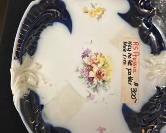 RS Prussia Blue Trim Porcelain Plate — This RS Prussia porcelain plate features a striking blue trim with ornate relief designs and delicate floral patterns in the center. The piece is an elegant example of early 20th century European porcelain craftsmanship. Measuring approximately 10 inches in length and 7.5 inches in width, it is suitable for display or collection. The plate is in good condition with no visible cracks but shows minor age-related wear. Originally made in Prussia, this collectible plate adds vintage charm to any collection.