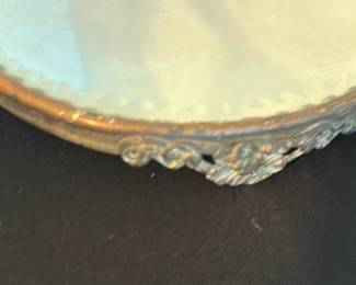 Ornate Round Vanity Table Mirror — This ornate round vanity table mirror features a decorative metal frame with intricate scrollwork and floral designs. The mirror glass is slightly aged, adding subtle vintage character suitable for a master bedroom setting. Measuring approximately 7 inches in diameter, it is a charming accent piece with a sturdy base and elegant detailing. The condition is good with minor wear consistent with gentle use and age. A perfect addition for vintage or antique-inspired decor collections.