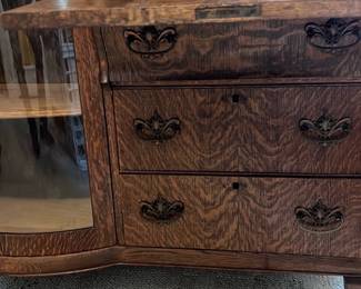 Antique Oak Secretary Curio Cabinet — 41”x68”. This antique oak secretary curio cabinet features intricate carved details and a glass display case. The piece combines functional storage with elegant design, including a fold-down writing surface, small drawers, and cubbies, topped with a decorative mirror and ornate carvings on the crest. Measuring approximately 59 inches tall, 43 inches wide, and 18 inches deep, it offers versatile use for office or living space. The cabinet is in good condition with typical minor wear consistent with age, showcasing the natural oak grain and craftsmanship. This classic piece represents early 20th-century furniture making with quality solid oak construction and distinctive embellishments.