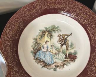 Vintage Victorian Scene Decorative Plate — This vintage decorative plate features a charming Victorian-era scene of two elegantly dressed ladies and a gentleman playing a lute. The plate showcases a rich burgundy border with intricate gold scrollwork that complements the detailed central illustration. Measuring approximately 10 inches in diameter, this plate serves as a stylish accent piece for any bedroom or display. It is in good condition with minor surface wear consistent with age and careful use. This collectible piece adds an elegant touch of historical charm to home decor.