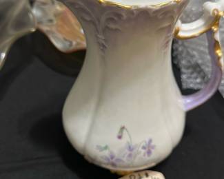 JP Vase Paris Porcelain Chocolate Pot — This delicate chocolate pot from JP Vase Paris is a beautifully crafted porcelain piece featuring elegant floral hand-painted designs in soft purples and greens. The pot has a gracefully curved form with ornate gold trim on the lid, spout, and handle, reflecting a refined vintage aesthetic. It measures approximately 7.5 inches in height, 5 inches in width, and 4.5 inches in length, making it a charming addition to any bedroom or display. The pot is in good vintage condition with minor signs of wear and no visible chips or cracks. This collectible porcelain piece bears a clear JP Vase Paris mark on the bottom, attesting to its authenticity and quality.