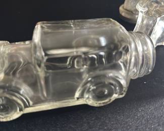 Vintage Glass Car Shaped Bottle — This vintage glass bottle is crafted in the shape of a classic car, featuring detailed embossed wheels and body contours. The clear glass construction highlights the charming automotive design, ideal as a collectible or decorative piece. The bottle measures approximately 4 in height, 2.5 in width, and 7 in length, making it a compact and eye-catching display item. It is in good condition with minor surface wear consistent with age and use. This unique car-shaped bottle adds character to any vintage collection or shelf display.