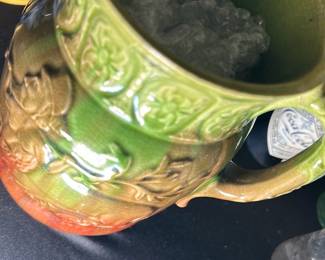 Green and Orange Ceramic Pitcher — This charming ceramic pitcher features a detailed relief design depicting a man playing a horn. The pitcher is glazed in vibrant green at the top, blending into warm orange at the base, with an intricate floral pattern adorning the rim. It measures approximately 8 inches in height and 6 inches in width, ideal for decorative display or functional use. The piece is in good condition with minor wear consistent with age, and no visible cracks or chips. Its unique design and warm colors make it an attractive addition to any vintage or rustic collection.