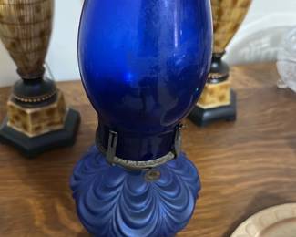Vintage Blue Glass Oil Lamp — This vintage oil lamp features a stunning cobalt blue glass chimney and reservoir with a decorative swirl pattern. The lamp includes an ornate metal base with intricate detailing, adding to its antique charm. Standing approximately 15 inches tall and 6 inches wide, it makes a beautiful accent piece for any bedroom or living space. The lamp is in good condition with some minor wear consistent with age, but the glass and metal components are intact. Its classic design evokes a nostalgic ambiance ideal for collectors or vintage decor enthusiasts.