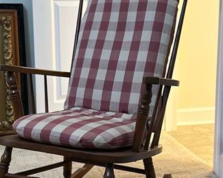 Wooden Rocking Chair with Cushions — This wooden rocking chair features a classic design with turned spindles on the arms and legs, paired with removable cushions in a red and white checkered pattern. The wood has a warm medium brown finish that complements the cozy fabric. Measuring approximately 40 in height, 25 in width, and 38 in depth, it is a comfortable size for relaxation in any room. The chair is in good condition with minor surface wear consistent with its usage. This traditional style rocking chair adds a charming touch to bedroom or living spaces.