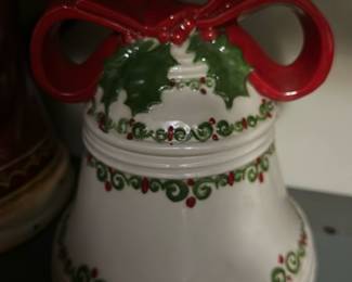 Ceramic Red Bow Christmas Bell — Festive ceramic bell with a large red bow and holly motif, perfect for holiday display. The white glazed bell features hand-painted green scrolls and red berry accents around the body and rim, with a prominent red ribbon handle on top. Measures approximately 7.5 inches tall and 5.5 inches wide, a lightweight decorative piece for a mantel or table. Good condition with minor surface wear consistent with display use and no visible chips or cracks.